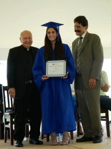 A smiling young lady in a blue cap and gown holding her diploma with a priest on one side and a man standing on her other side