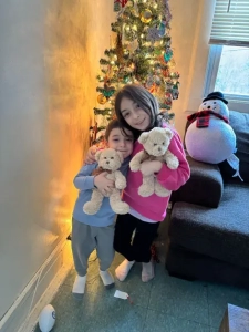 A girl with her arm around a young boy both holding teddy bears and standing in front of a Christmas tree