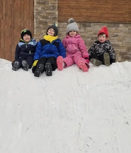 Four children in winter gear sitting on the snow at the top of a slope