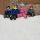 Four children in winter gear sitting on the snow at the top of a slope