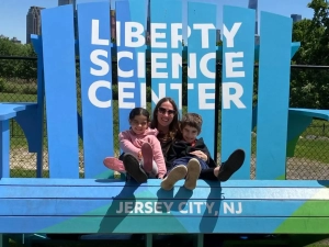 A woman with a girl and boy sitting on her lap in a giant blue Adirondack chair with "Liberty Science Center" on the back