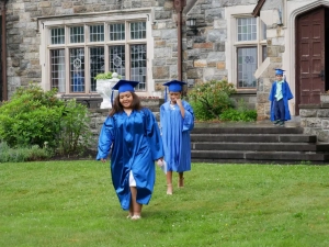 Two Saint Basil graduates dressed in cap and gown walking in the grass in front of Saint Basil Academy's main building.