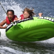 Two children in life jackets hanging on to a bright green inflatable inner tube being pulled on the water.