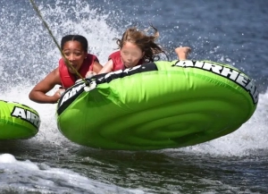 Two children in life jackets hanging on to a bright green inflatable inner tube being pulled on the water.