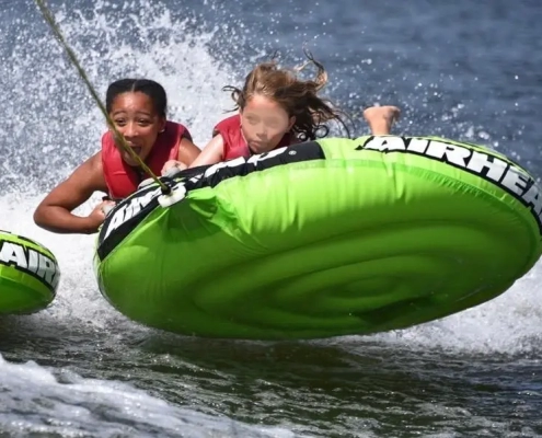 Two children in life jackets hanging on to a bright green inflatable inner tube being pulled on the water.