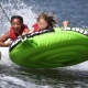 Two children in life jackets hanging on to a bright green inflatable inner tube being pulled on the water.