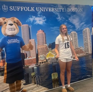 A young lady from Saint Basil Academy holding a lacrosse stick and standing in front of a Suffolk University backdrop