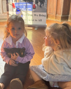 A little girl holding a young black chicken while sitting on the floor next to another little girl