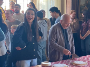 A lady smiling standing next to Father Costa about to cut Vasilopita and a number of people in the background