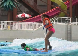 A girl standing on a surf board in the wave pool at a surf park and next to her is a boy laying down on a surfboard.