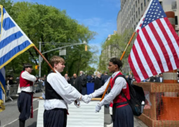 Two young men stand facing each other shaking hands. The youth on the left has a Greek flag on a pole resting on his shoulder and the youth on the right has an American flag.