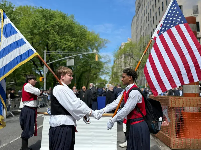 Two young men stand facing each other shaking hands. The youth on the left has a Greek flag on a pole resting on his shoulder and the youth on the right has an American flag.