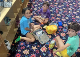 2026 Summer activities Three Saint Basil Academy children sitting on a colorful carpet eating their lunch.