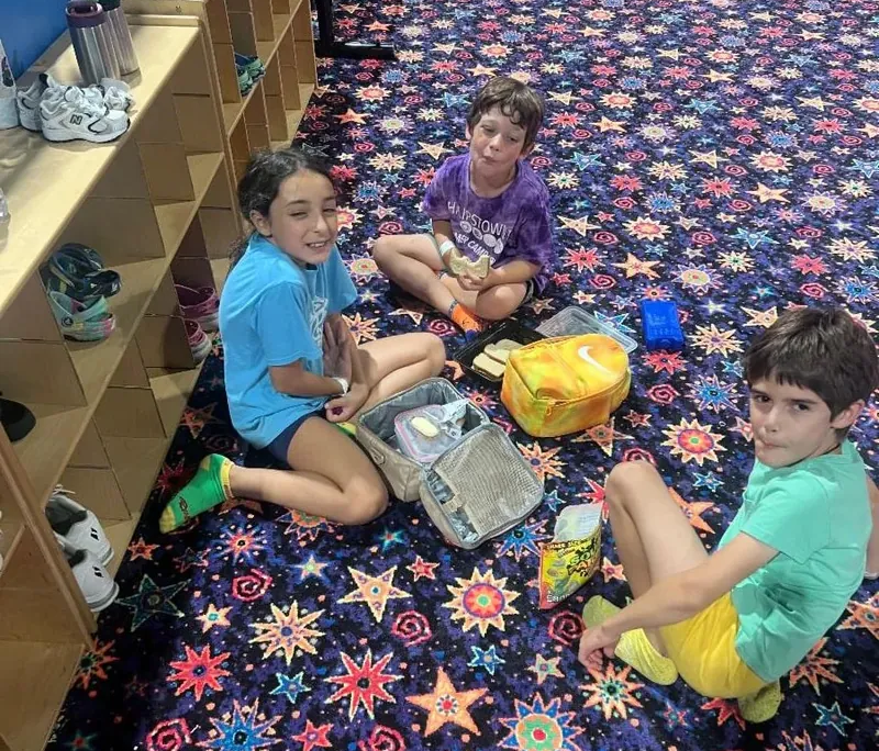 2026 Summer activities Three Saint Basil Academy children sitting on a colorful carpet eating their lunch.
