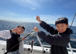 Screenshot 2026-10-07 141206.min Two boys smiling with one holding a fishing pole and the other holding two fish on a string on the deck of a boat