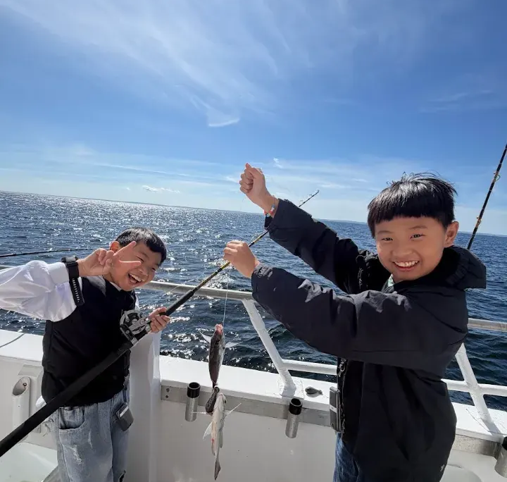 Screenshot 2026-10-07 141206.min Two boys smiling with one holding a fishing pole and the other holding two fish on a string on the deck of a boat