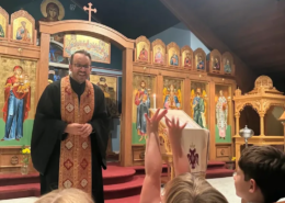 St. Anastasia Paraklesis Father Nikolas Delaveris smiling as he is talking at the front of the Saint Basil Chapel with children in the pews