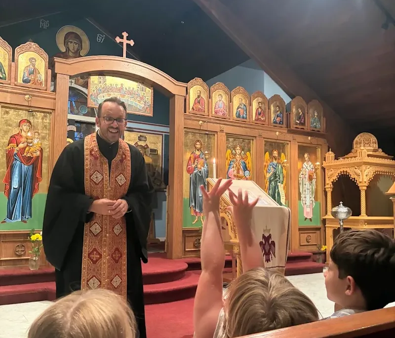 St. Anastasia Paraklesis Father Nikolas Delaveris smiling as he is talking at the front of the Saint Basil Chapel with children in the pews